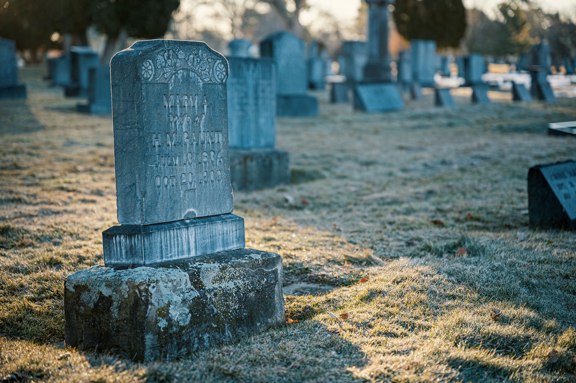A headstone standing in frost, with a cemetery behind it